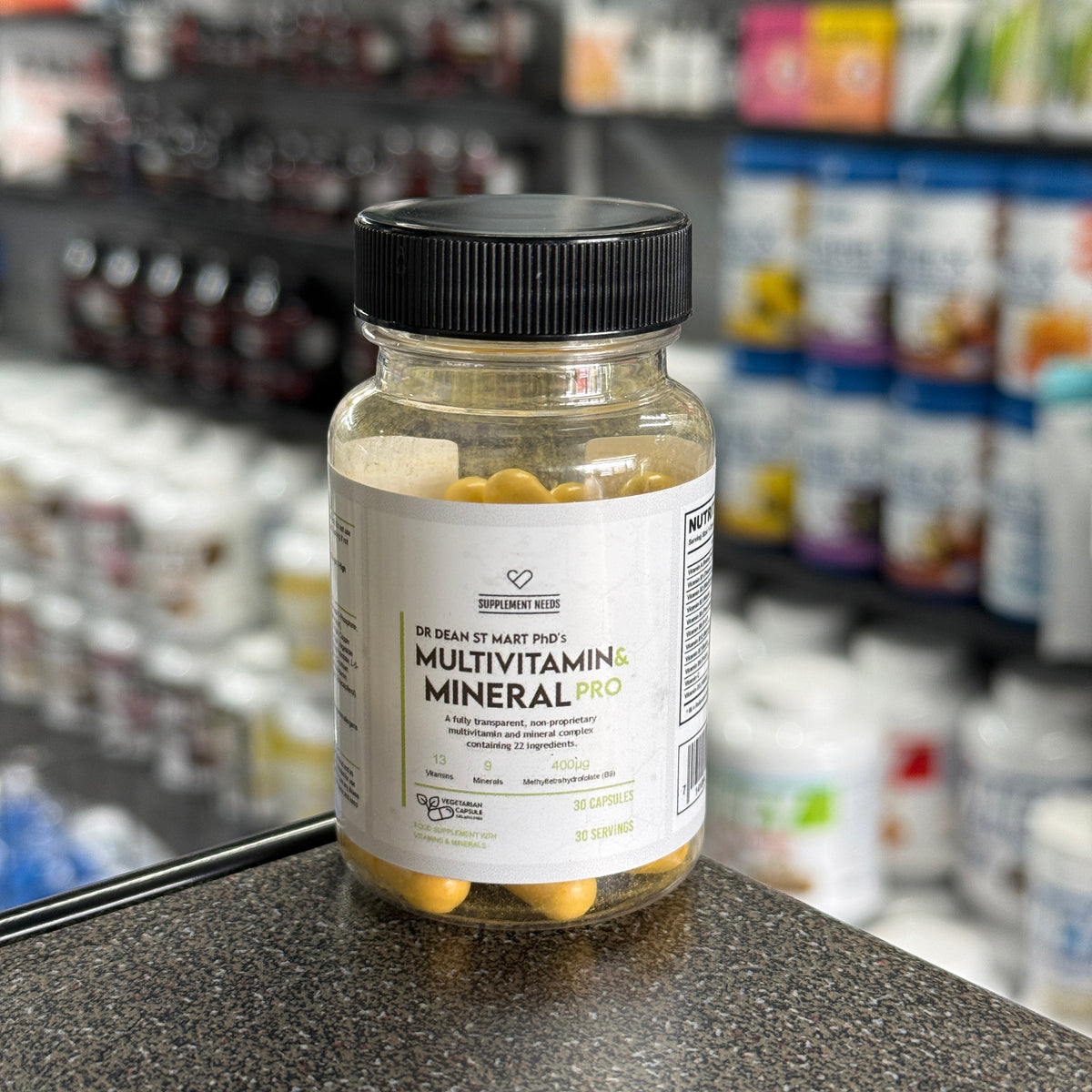 A bottle of Dr Dean St Mart's Multivitamin & Mineral Pro dietary supplement, containing 30 capsules, placed on a dark countertop with a blurred supplement store background.
