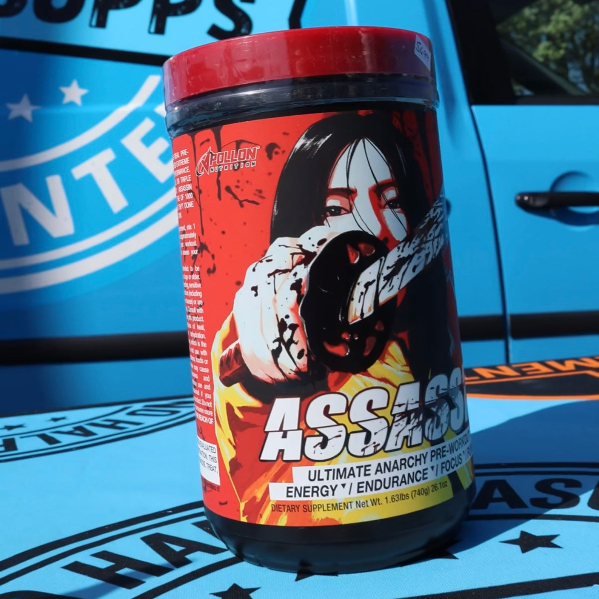 A tub of Assassin Extreme pre-workout supplement by Apollon Supplements, featuring a red lid and a red, black, and yellow label with a graphic of a woman holding a gun, set against a blue vehicle background.