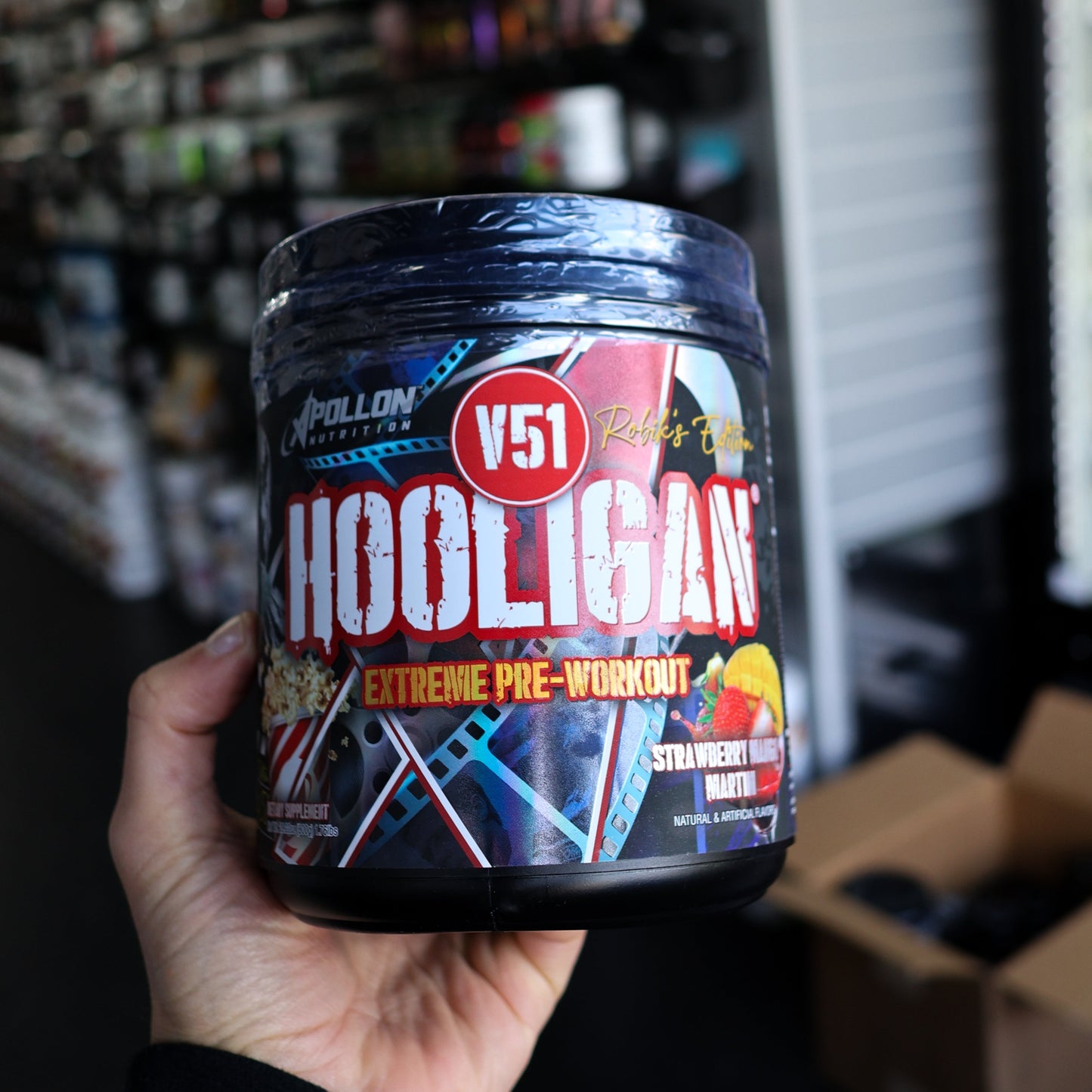 Container of V51 Hooligan pre-workout supplement held in a hand with a store shelf in the background.