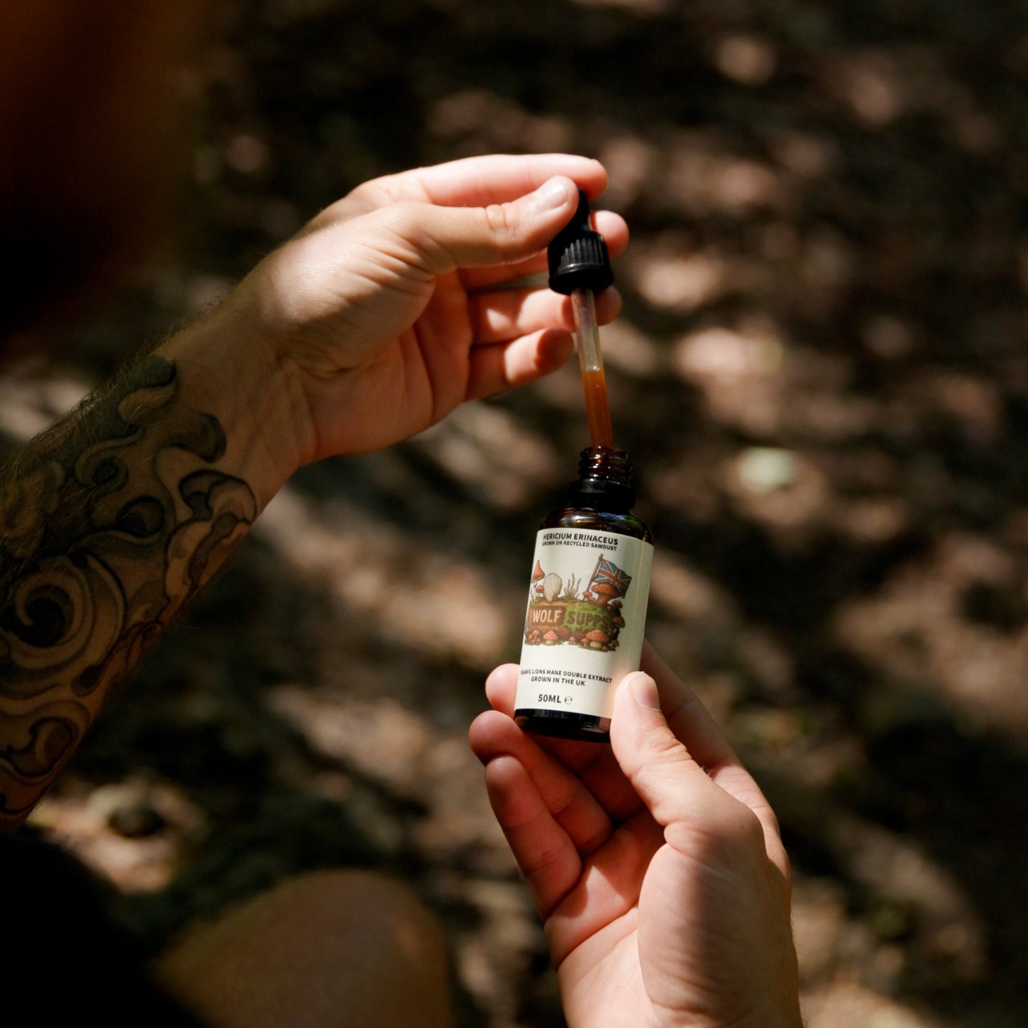 A person with tattooed arms holds a small amber dropper bottle outdoors in dappled sunlight, with the dropper poised above the bottle.