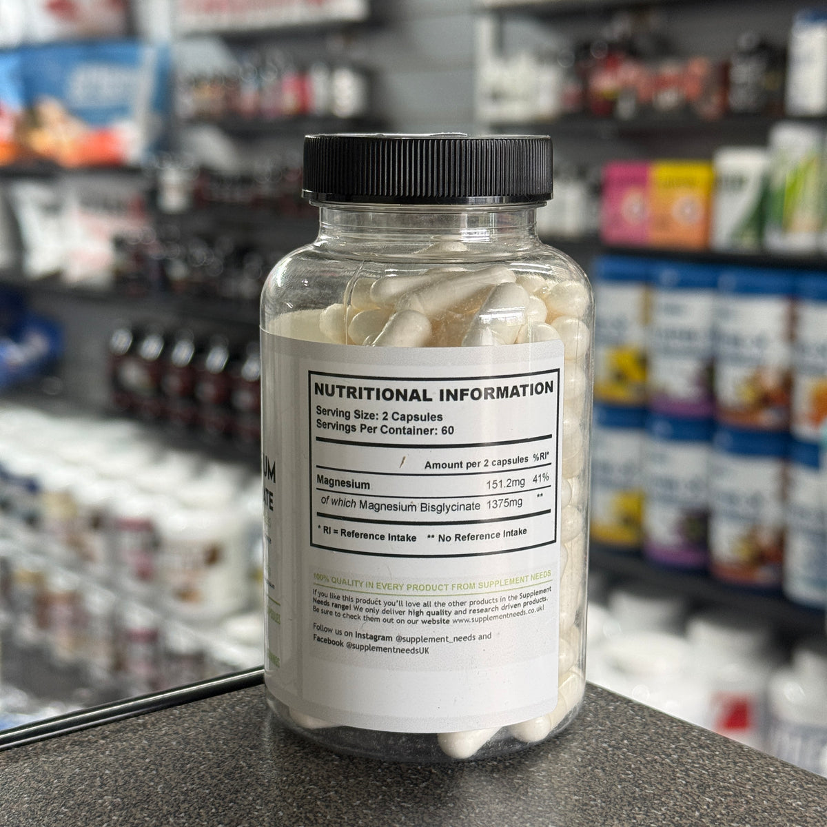 A clear supplement bottle filled with white capsules, featuring a black cap and a white label with a nutrition facts panel, set on a countertop with a blurred store background.