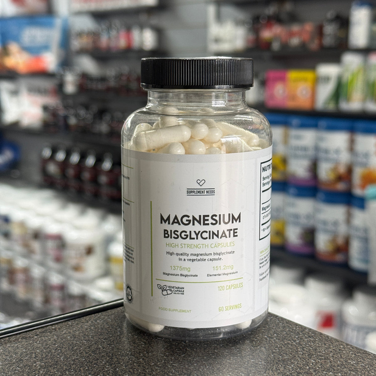 A bottle of Supplement Needs Magnesium Bisglycinate dietary supplement capsules, containing 120 capsules, set on a countertop with a blurred supplement store background.