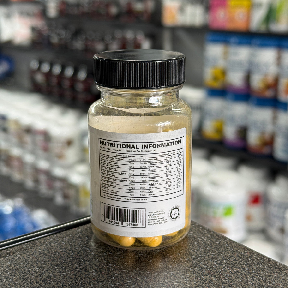 A clear supplement bottle with a black ribbed cap, showing the back label with nutritional information and yellow tablets inside, set on a countertop with a blurred supplement shop background.