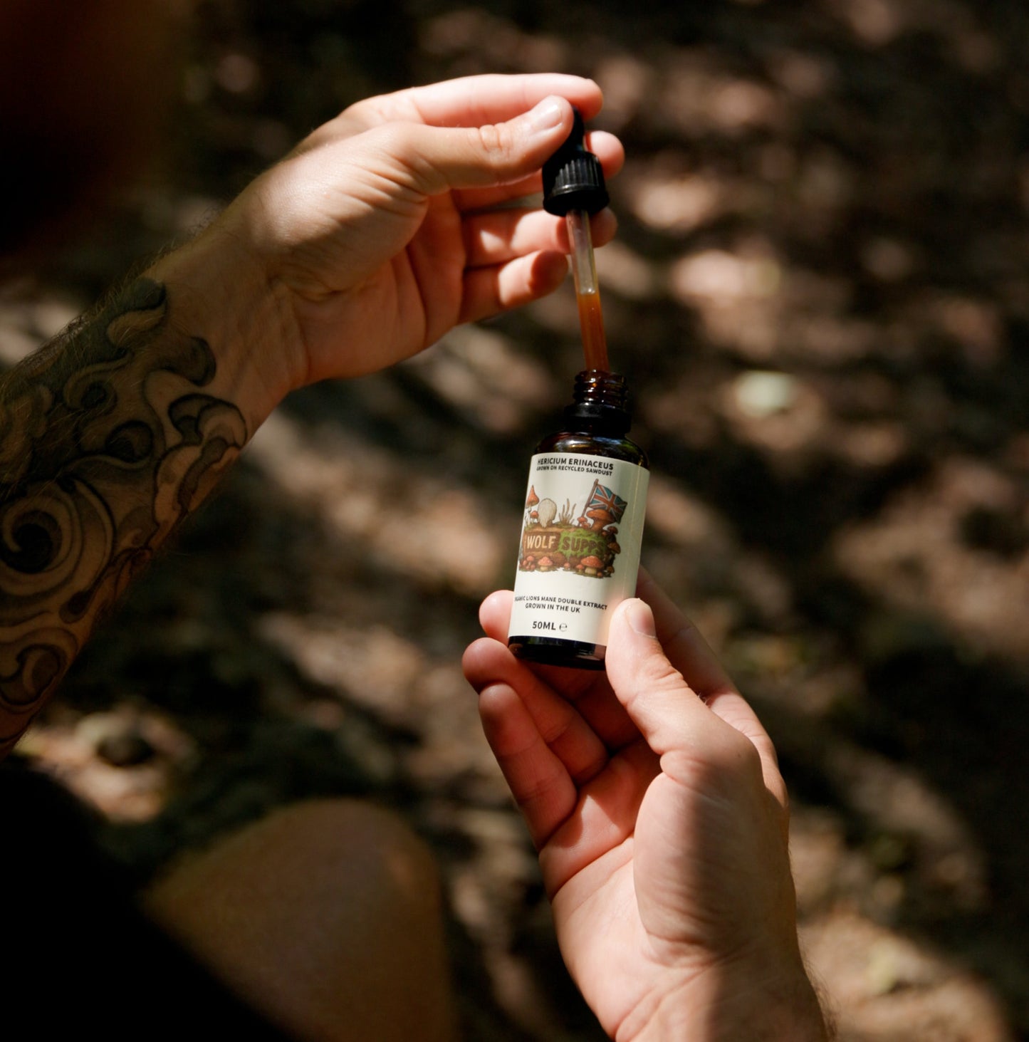 A person with tattooed arms holds a small amber dropper bottle outdoors in dappled sunlight, with the dropper poised above the bottle.
