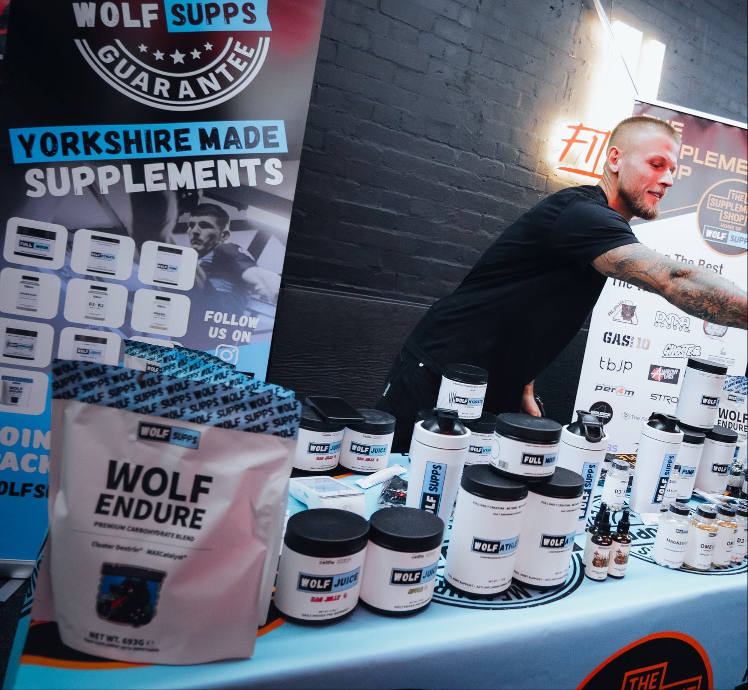 A product display booth for Wolf Measures Supplements, featuring a large 'Wolf Endure' bag, multiple white supplement tubs with black lids, and branded roll-up banners. A man is visible behind the table against a dark brick wall background.